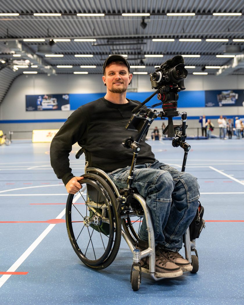 Johannes Springer in seinem Rollstuhl in einer Sporthalle, mit einer Profikamera auf einem selbst gebauten Haltesystem.
