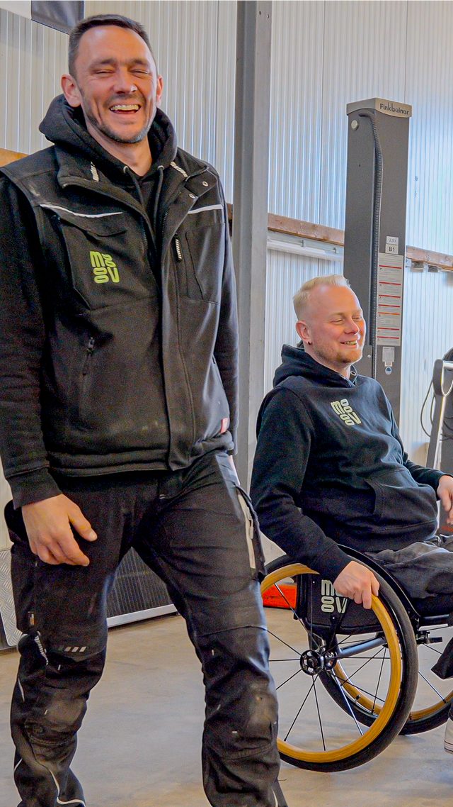 Standbild aus einem Social-Clip: zwei Mitarbeiter in Arbeitskleidung lachen in einer Werkstatt, einer von ihnen sitzt im Rollstuhl.
